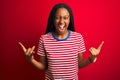 Young african american woman wearing striped t-shirt standing over isolated red background shouting with crazy expression doing Royalty Free Stock Photo