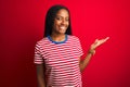 Young african american woman wearing striped t-shirt standing over isolated red background smiling cheerful presenting and Royalty Free Stock Photo