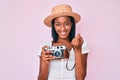 Young african american woman wearing summer hat holding vintage camera beckoning come here gesture with hand inviting welcoming