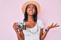 Young african american woman wearing summer hat holding vintage camera crazy and mad shouting and yelling with aggressive Royalty Free Stock Photo