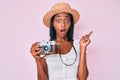 Young african american woman wearing summer hat holding vintage camera surprised pointing with finger to the side, open mouth Royalty Free Stock Photo