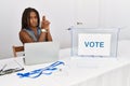 Young african american woman working at political election sitting by ballot holding symbolic gun with hand gesture, playing