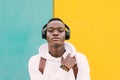 Young african black man against a yellow and green wall wearing a white sweatshirt and a backpack listening music on wireless head Royalty Free Stock Photo