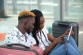 Young african couple checking their travel schedule on a tablet, sitting at the station with their suitcases