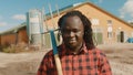 Young african farmerholding fork over the soulder and smiling in front of the silo system