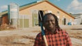 Young african farmerholding fork over the soulder and smiling in front of the silo system