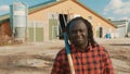 Young african farmerholding fork over the soulder and smiling in front of the silo system