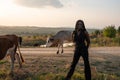 Young african girl in black clothes stands among the field, holds her hands behind her back, cows graze on the