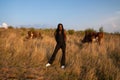 Young african girl in black clothes stands among the field, holds her hands behind her back, cows graze on the