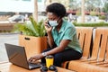 A young African girl in a medical face mask is talking on the phone and working with a laptop at a table in a cafe Royalty Free Stock Photo