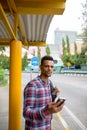 Young African hipster man with backpack using phone while waiting at the bus stop