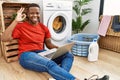 Young african man doing laundry and using computer smiling positive doing ok sign with hand and fingers Royalty Free Stock Photo