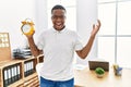 Young african man holding alarm clock at the office celebrating victory with happy smile and winner expression with raised hands Royalty Free Stock Photo