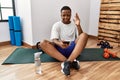 Young african man sitting on training mat at the gym using smartphone gesturing finger crossed smiling with hope and eyes closed Royalty Free Stock Photo