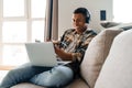 Young african man studying on laptop and writing down notes while sitting on couch Royalty Free Stock Photo