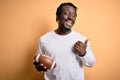 Young african player man playing rugby holding american football ball over yellow background pointing and showing with thumb up to Royalty Free Stock Photo