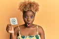 Young african woman with afro hair holding online mail symbol on paper scared and amazed with open mouth for surprise, disbelief