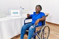 Young african woman sitting on wheelchair voting putting envelop in ballot box looking at the camera blowing a kiss on air being