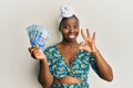 Young african woman wearing hair turban holding south african 100 rand banknotes doing ok sign with fingers, smiling friendly