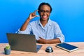 Young african woman working at hotel reception using laptop smiling and confident gesturing with hand doing small size sign with Royalty Free Stock Photo