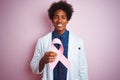 Young afro american doctor man holding cancer ribbon standing over isolated pink background with a happy face standing and smiling Royalty Free Stock Photo