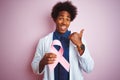 Young afro american doctor man holding cancer ribbon standing over isolated pink background pointing and showing with thumb up to Royalty Free Stock Photo