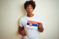 Young afro american man holding Serbia Serbian flag standing over isolated white background scared in shock with a surprise face, Royalty Free Stock Photo