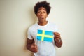 Young afro american man holding Sweden Swedish flag standing over isolated white background scared in shock with a surprise face, Royalty Free Stock Photo