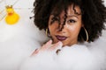 Young afro-american teen girl laying in bath with foam, wearing swag jewelry flawless, making selfie Royalty Free Stock Photo