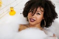 Young afro-american teen girl laying in bath with foam, wearing swag jewelry flawless, making selfie Royalty Free Stock Photo