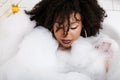 Young afro-american teen girl laying in bath with foam, wearing swag jewelry flawless, making selfie Royalty Free Stock Photo