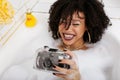 Young afro-american teen girl laying in bath with foam, wearing swag jewelry flawless, making selfie Royalty Free Stock Photo