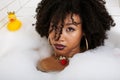 Young afro-american teen girl laying in bath with foam, wearing swag jewelry flawless, making selfie Royalty Free Stock Photo