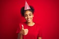 Young afro american woman wearing birthday hat over isolated red background doing happy thumbs up gesture with hand