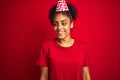 Young afro american woman wearing birthday hat over isolated red background looking away to side with smile on face, natural Royalty Free Stock Photo