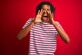 Young afro man with dreadlocks wearing striped t-shirt standing over isolated red background Shouting angry out loud with hands Royalty Free Stock Photo