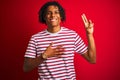 Young afro man with dreadlocks wearing striped t-shirt standing over isolated red background smiling swearing with hand on chest Royalty Free Stock Photo