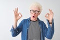 Young albino blond man wearing denim shirt and glasses over isolated white background looking surprised and shocked doing ok