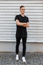 Young amazing stylish man in a black stylish t-shirt in trendy jeans in white sneakers poses near a white wooden vintage house. Royalty Free Stock Photo