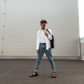 Young American fashion model man in a vintage white shirt in blue stylish jeans with a black cloth bag poses in the city on a Royalty Free Stock Photo