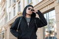 Young American hipster man in a stylish hooded sweatshirt in dark sunglasses in a cap with a backpack is standing in a city Royalty Free Stock Photo