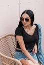 Young American woman with long hair in sunglasses in a stylish denim jacket in a black t-shirt sitting on a chair near a pink wall Royalty Free Stock Photo