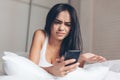 Young Annoyed angry woman looking on mobile phone in bed. Smiling Brunette Girl holding mobile phone