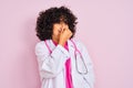 Young arab doctor woman with curly hair wearing stethoscope over isolated pink background smelling something stinky and