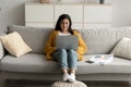 Young arab female freelancer using laptop wearing wireless earphones and typing on keyboard, sitting on sofa at home Royalty Free Stock Photo