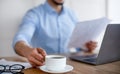 Young Arab guy reading documents, using laptop, drinking coffee while working at office, closeup Royalty Free Stock Photo