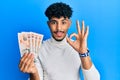 Young arab handsome man holding 10 united kingdom pounds banknotes doing ok sign with fingers, smiling friendly gesturing