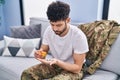 Young arab man army soldier taking pills sitting on sofa at home