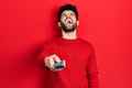 Young arab man with beard holding television remote control angry and mad screaming frustrated and furious, shouting with anger Royalty Free Stock Photo