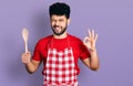 Young arab man with beard wearing baker uniform holding wooden spoon winking looking at the camera with sexy expression, cheerful Royalty Free Stock Photo